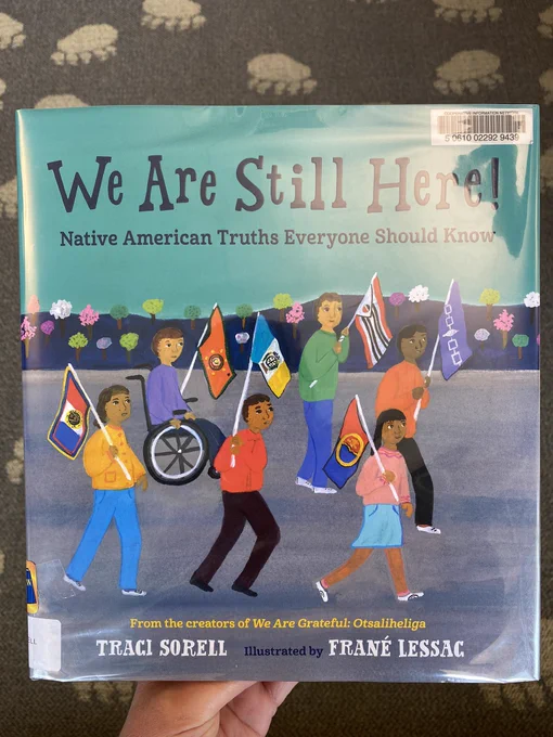 Mandi holds a library copy of We Are Still Here! Native American Truths Everyone Should Know, anonfiction picture book written by Traci Sorell and illustrated by Frané Lessac.