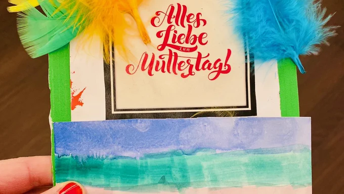 Auf dem Foto hält eine Hand zwei von Kindern gebastelte Karten. Auf der unteren sind Streifen in Regenbogenfarben, die obere ist mit bunten Federn geschmückt, in der Mitte steht: "Alles Liebe zum Muttertag!"