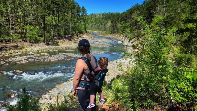 Amy stands by a large river in the forest while carrying her son.