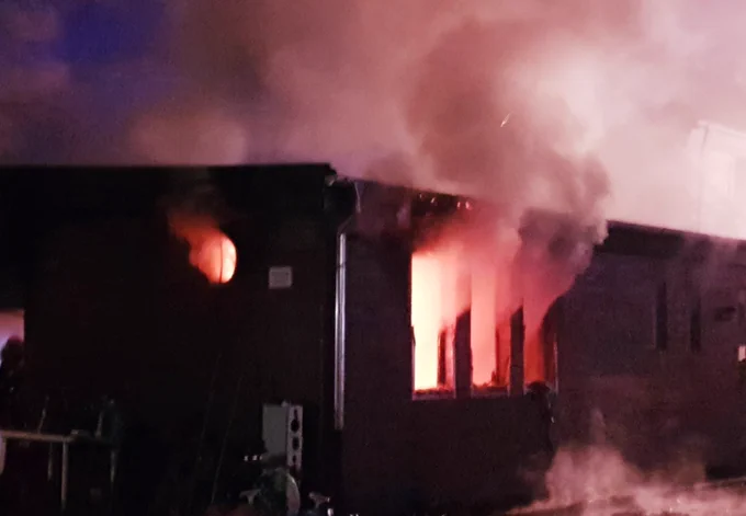 Ein flaches Haus. Aus den Fenstern dringt Rauch, im Inneren sieht man rot leuchtend Feuer.