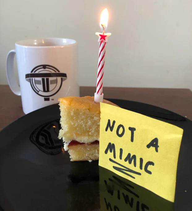A piece of birthday cake with a lit red candle sits on a plate with a 'not a Mimic' sticky-note attached, with a mug of tea featuring the Transtar logo in the background