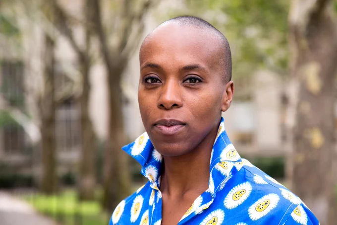 African woman in brightly colored blouse smiling slightly at the camera.