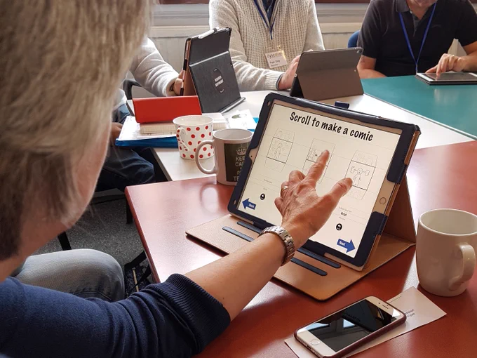 Photograph - an over-the-shoulder view of a person touching an iPad screen entitled "Scroll to make a comic". They are seated at a table alongside other people working at iPads
