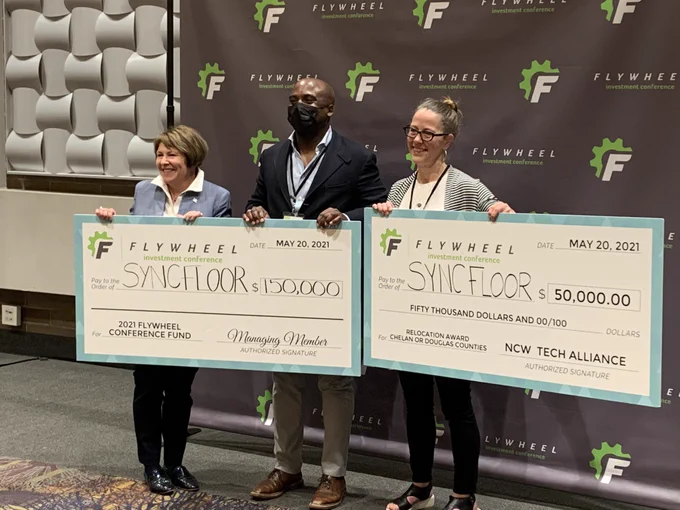 Lisa Brown is standing next to the two leaders of Syncfloor. They are each holding a corner of two large checks showing their award amounts of $150,000 and relocation award of $50,000. They are standing in front of a dark brown backdrop that shows the Flywheel logo and they are smiling at a camera.