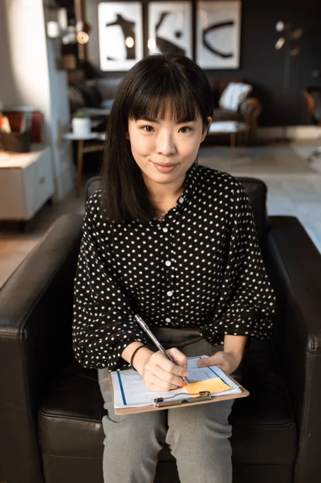 Woman wearing a black and white polka dot shirt sitting on a black leather seat holding a clipboard and pen.