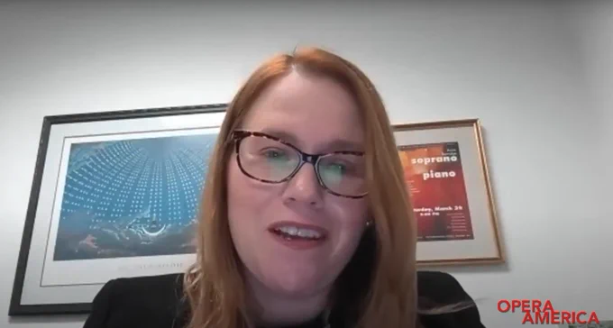 Annie Burridge on web cam, smiling at us as she talks, in front of a white wall and two artworks. The OPERA America logo is bottom right of the screen.