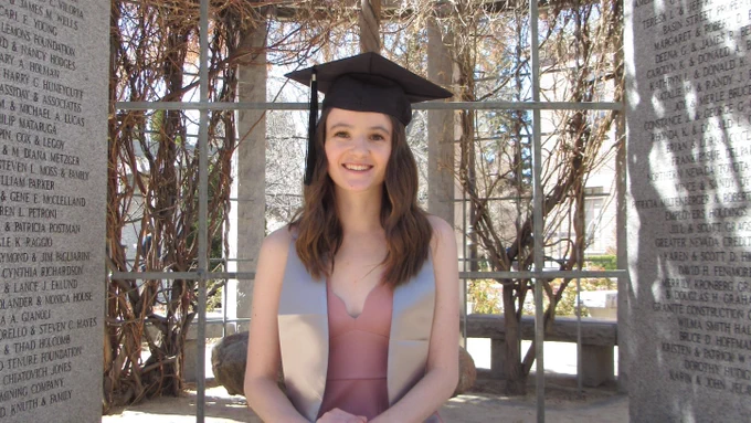 Student writer Jordan Nicholson sits on a bench between two granite walls wearing her graduation cap.