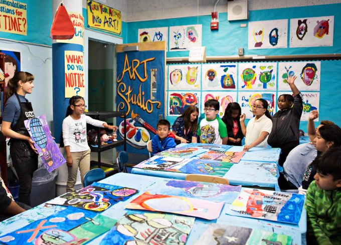 a group of kids in an art classroom which is filled with all sorts of colorful work on paper