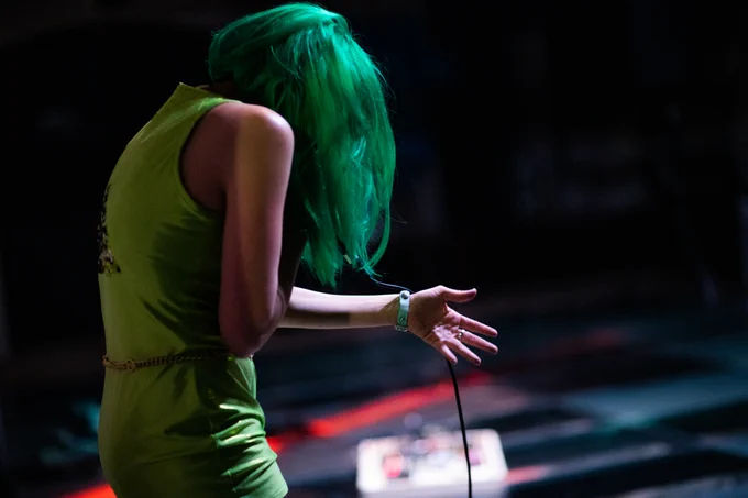 Photo of Leyya in profile against a dark background. She wears a green wig and clothes, holds a mic & extends her left hand