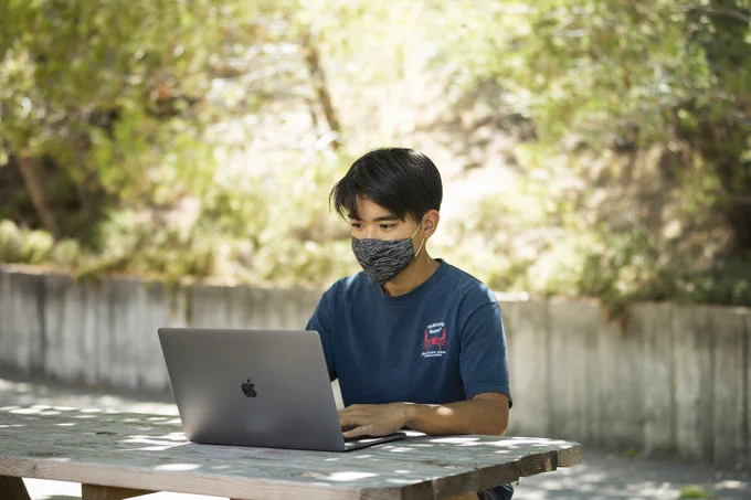 A young man wearing a cloth face mask sitting outdoors and typing on a laptop.
