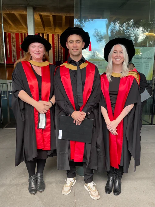 3 people in academic dress smiling for the camera. They are wearing black robes with red and gold sash while standing in front of glass doors.