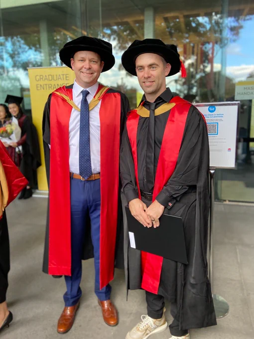 2 people in academic dress smiling for the camera. They are wearing black robes with red and gold sash while standing in front of glass doors.