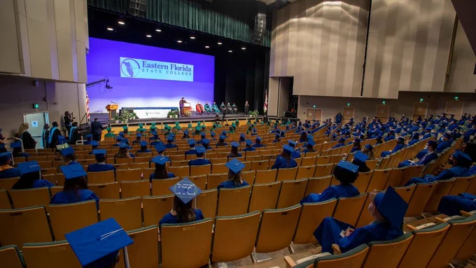 Socially distanced grads in blue and green caps and gowns sit in front of the King Center stage.