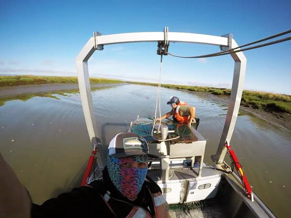 A selfie view of two people in a boat. One is wearing a cap and mask, looking up at the camera, while the other sits at the helm. Both are wearing life jackets as they navigate through a delta.