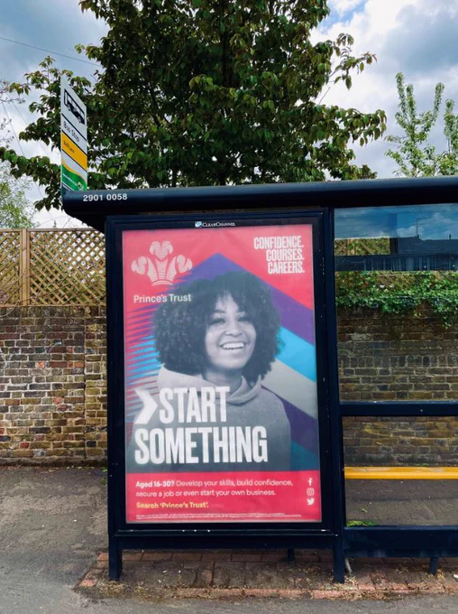 Start Something brand advert from Prince's Trust at bus stop. Features mixed race woman with curly hair wearing red hoodie, smiling. 