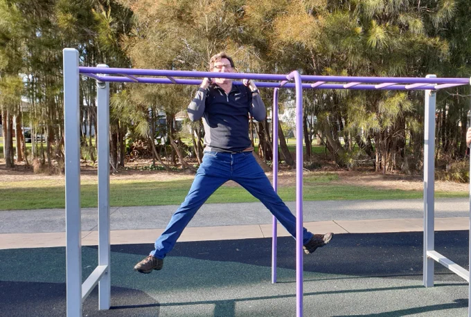 middle aged man with short brown hair wearing jeans and a long sleeved shirt swinging from monkey bars