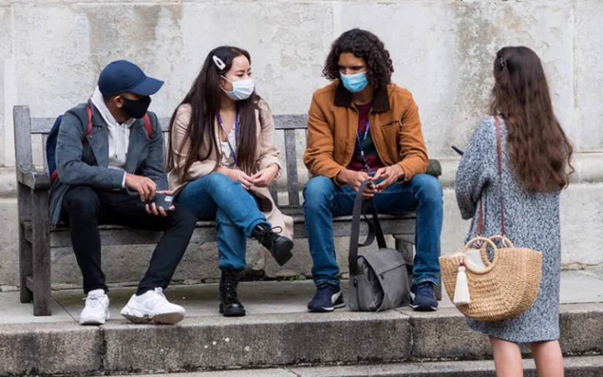 Three UCL students sit on a bench in the main quad wearing face coverings and talking. Another stands facing them.
