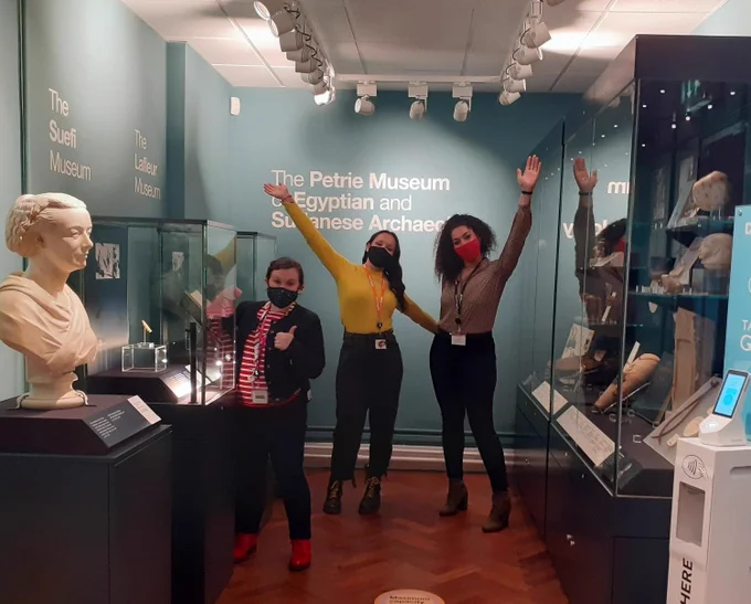 The main entrance gallery of the museum. A bust of Amelia Edwards and some glass museum cases are in the foreground. Three women stand underneath a sign showing the museum's name. They are wearing masks and waving. One is cleaning a museum case with duster. 