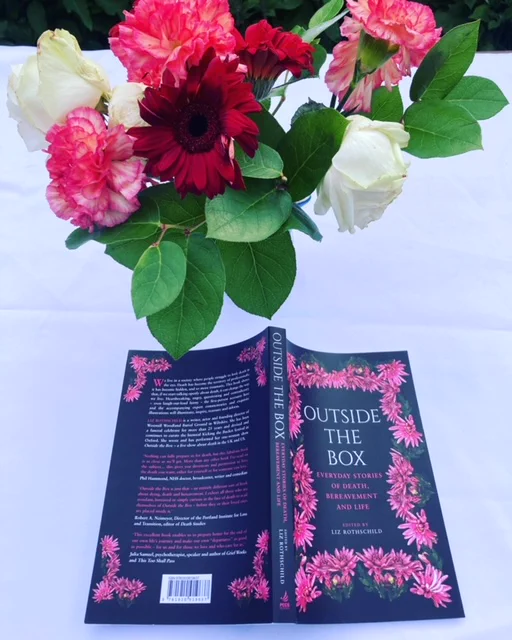 Book cover entitled Outside the box - everyday stories of life, death and bereavement by Liz Rothchild. The cover is black with white writing and bright pink flowers on it. There is also a vase of pink, white and red flowers next to the book.