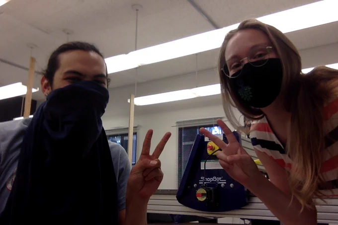 A masked man and woman making peace signs with their fingers standing in front of a CNC mill