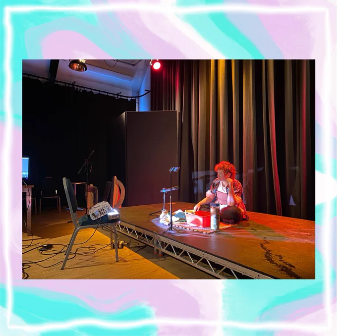 A person in a studio sat cross legged on a small black stage. They have a red tub and a tube on paper mulch in front of them on a small mat. There are stage lights and a projector lighting them with white, red and yellow light. There are black draped hanging from the studio walls.