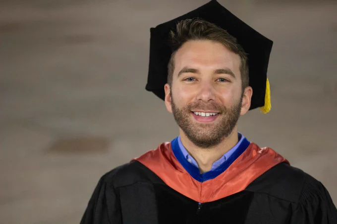 Medium close-up of a man in PhD graduation regalia, smiling.