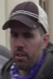 A man with a mustache and light beard wears a gray or black and white baseball cap with two white stripes across the front, and a dark colored jacket over a blue and white shirt or scarf.