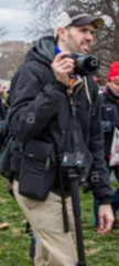 A man with a mustache and light beard wears a gray or black and white baseball cap with white stripes across the front, a black jacket, khaki pants, and a black bag that sits on his right hip. He is holding a black camera with a large lens in his right hand, and appears to have a black tripod in front of him.