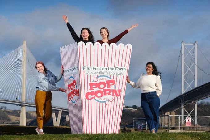 A large popcorn box sits next to the Forth, with the bridges in the background. Two young women stand in the box and two women stand either side smiling happily.