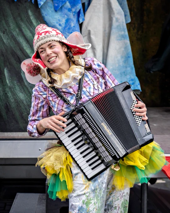 An actor dressed in a green and yellow tutu, checked shirt and comedic woolly hat plays the accordion and smiles broadly at the audience