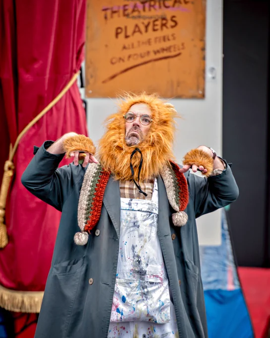 An actor wearing a lion's shaggy mane, furry fingerless gloves, a woolly scarf, overcoat and paint-splashed overalls looks out into the audience with a mock sad face