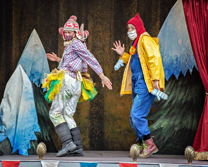 Two actors in colourful red, yellow, green and blue outfits dance energetically on the back of a truck decked out like a stage