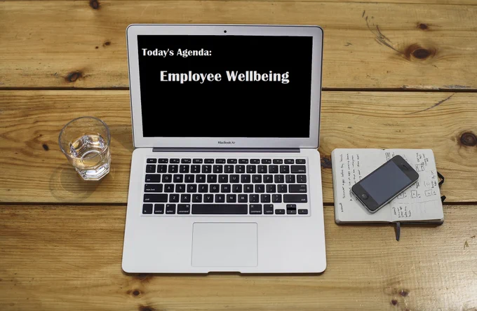Image of looking down at a wooden workstation with the following items (left to right) glass of water, laptop with the information: 'Today's Agenda; Employee Wellbeing' on the screen, and finally a mobile phone positioned on a notebook.