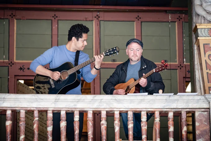 Two actors stand on a balcony, playing acoustic guitars.
