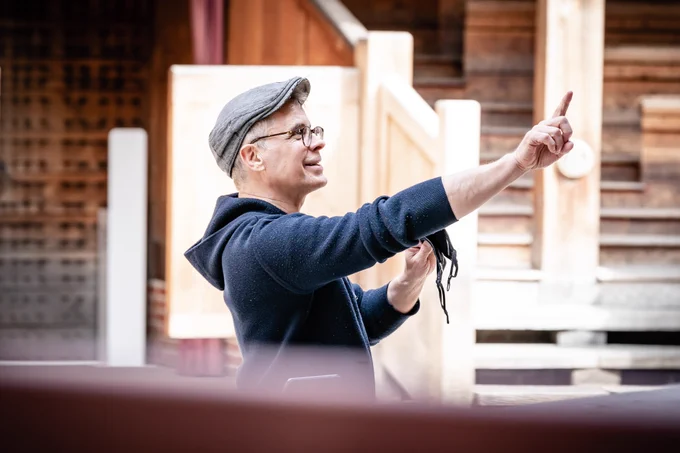 A Director wearing glasses and a peak flat cap smiles and raises their arms out, pointing with their hand.