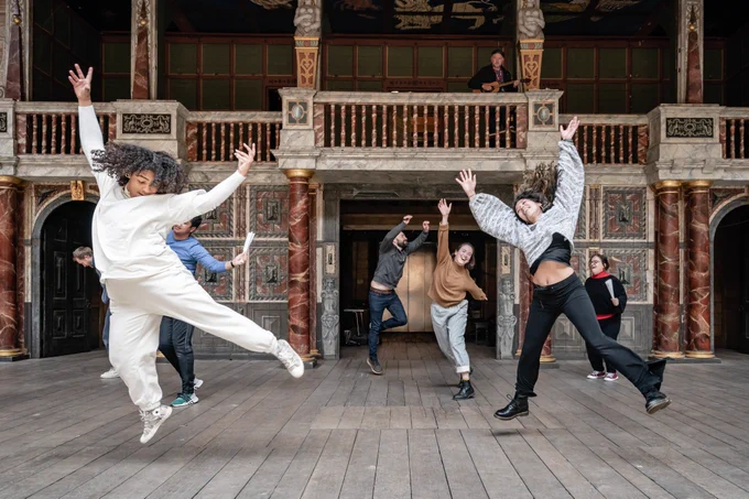A group of actors dance and leap into the air, their arms outstretched on the Globe Theatre stage.