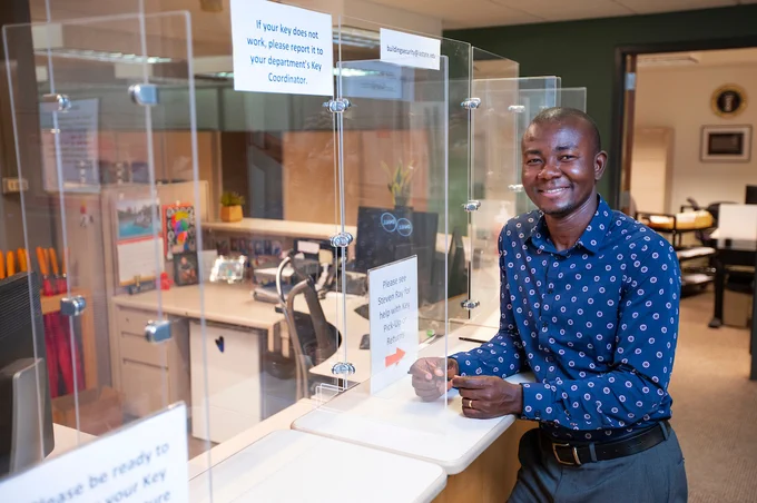 A man stands in front of plexiglass barriers at a desk