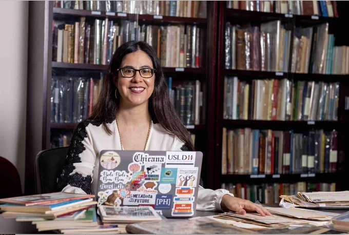 Woman seated at a table, behind a laptop and in front of bookcases. Historic documents on table.