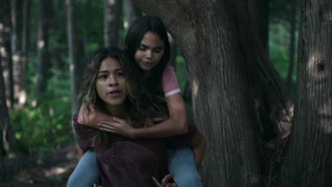 Gina Rodriguez carrying Ariana Greenblatt's character on her back in a woodsy area in "Awake"