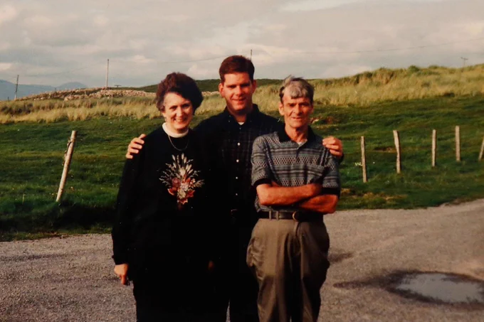 An old photo of Secretary Marty Walsh with his parents. They are standing on gravel with a hill full of green grass and weeds in the background. 