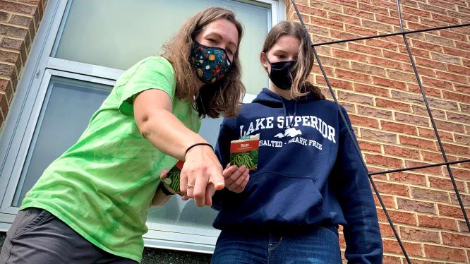 Teacher and student standing together outside by a brick wall. The teacher is pointing down and the student is holding a bag of bean seeds.