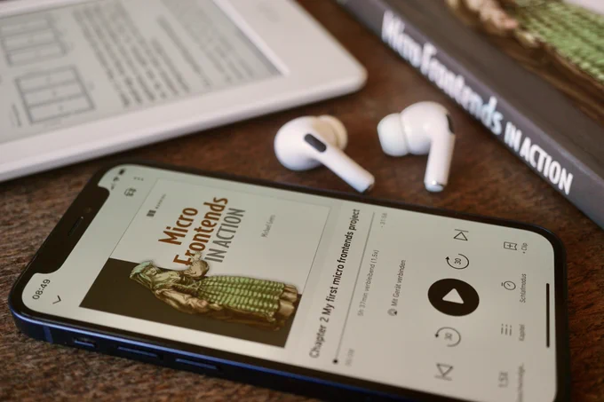 Phone and Headphones playing the Micro Frontends in Action audiobook. Paper and kindle version of the book are visible in the background.