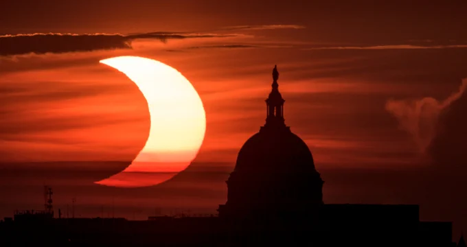 The Sun, about half-covered by the Moon, is visible in a sunrise sky behind a dome