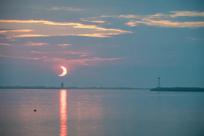 The Sun, about half-covered by the Moon, is visible at sunrise over the water