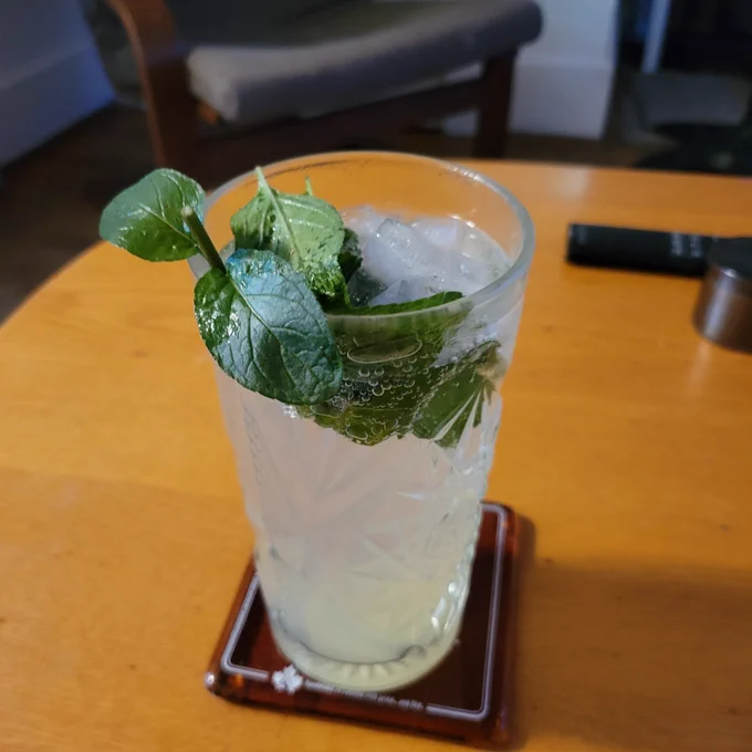 A tall glass sits on a coaster filled with ice, mint leaves and bubbly sweet mojito.