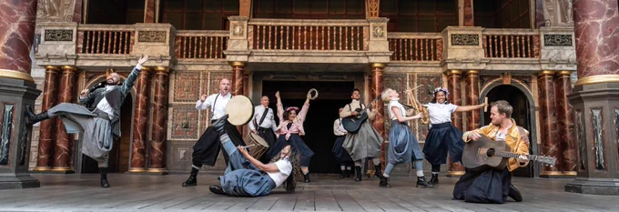 A group of actors dance, play instruments and raise their arms enthusiastically into the air on the wooden stage of the Globe Theatre.