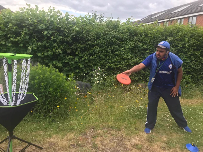 Young man in a garden with a hedge and some houses behind him. He is about to throw a frisbee into a basketball like net. He is wearing a  Chelsea football club hat and top, with dark blue tracksuit bottoms and bright blue trainers.
