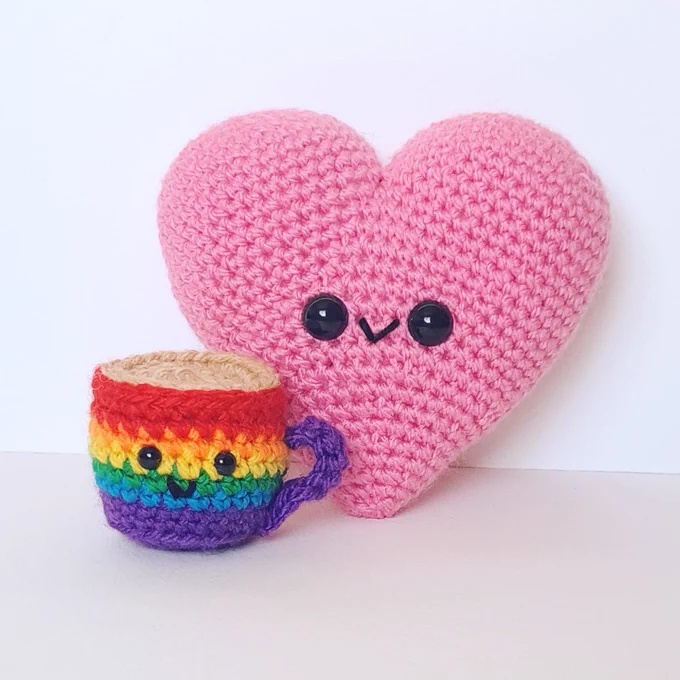 A crocheted heart with a crocheted coffee cup. The heart is pink and the coffee cup is rainbow striped. Both have a smiling face.