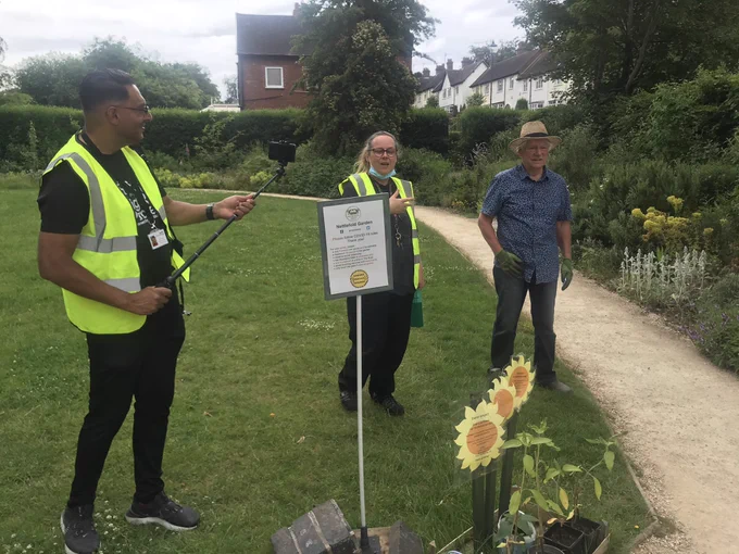 A man holding a camera films a man and woman walking in a sensory garden