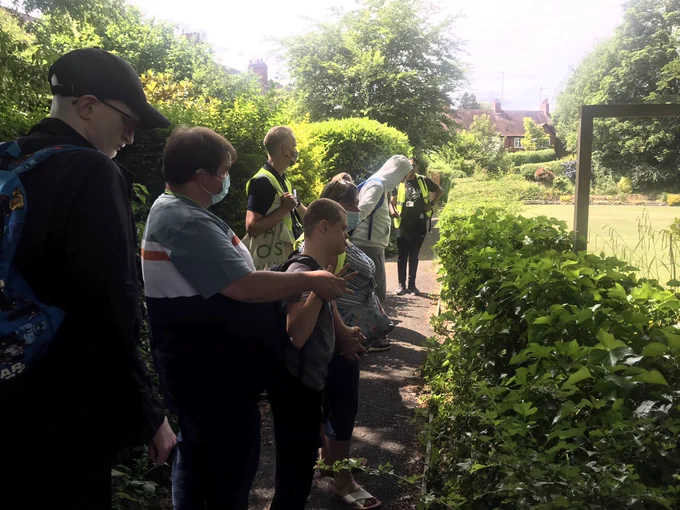 A group of people standing in front of a hedge look over at a bowling green